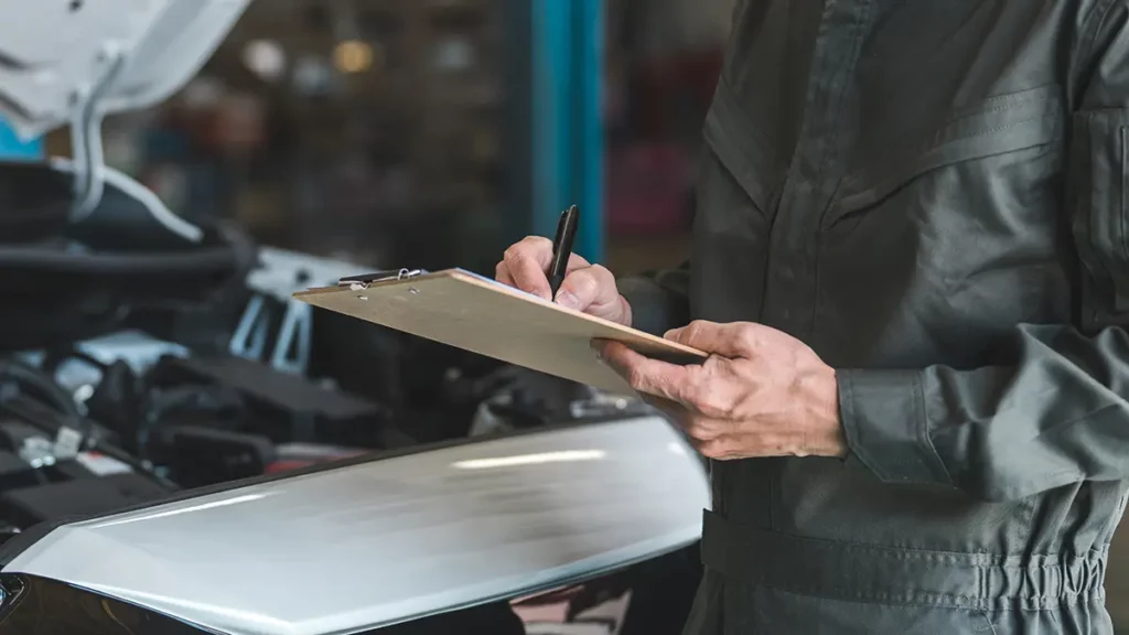 A mechanic wearing a uniform is holding a clipboard and writing notes while inspecting a vehicle's engine.